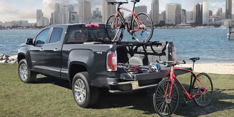 A GMC Canyon truck with one bicycle inside the truck bed and another bicycle placed outside on the ground next to it.